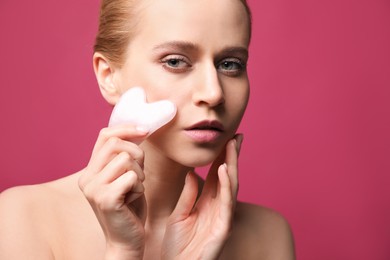 Photo of Beautiful young woman doing facial massage with gua sha tool on pink background, closeup