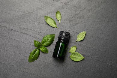 Photo of Bottle of essential basil oil and fresh leaves on dark grey table, flat lay