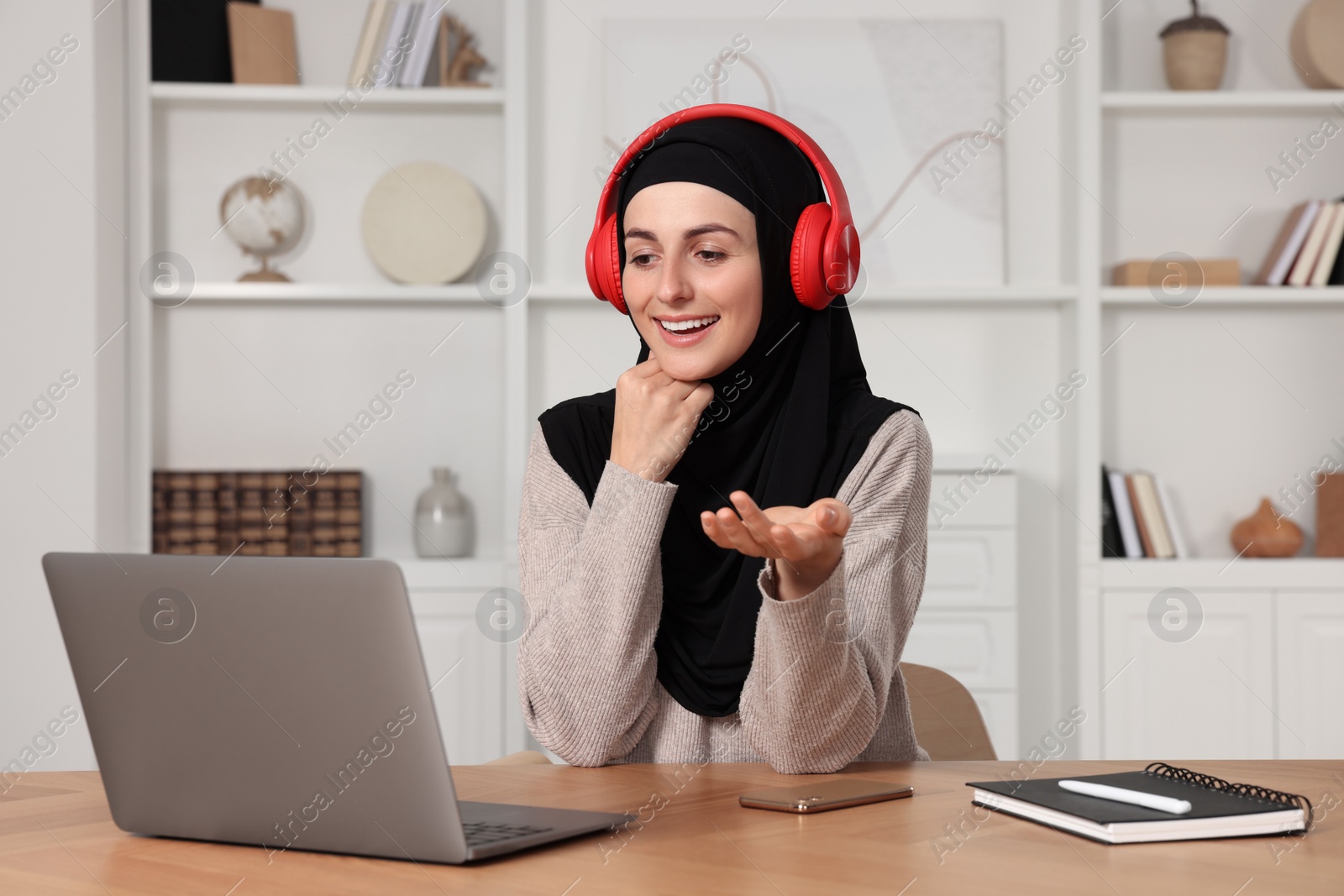 Photo of Muslim woman in hijab using video chat on laptop at wooden table in room