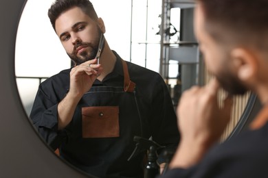 Photo of Handsome young man trimming beard with scissors near mirror indoors