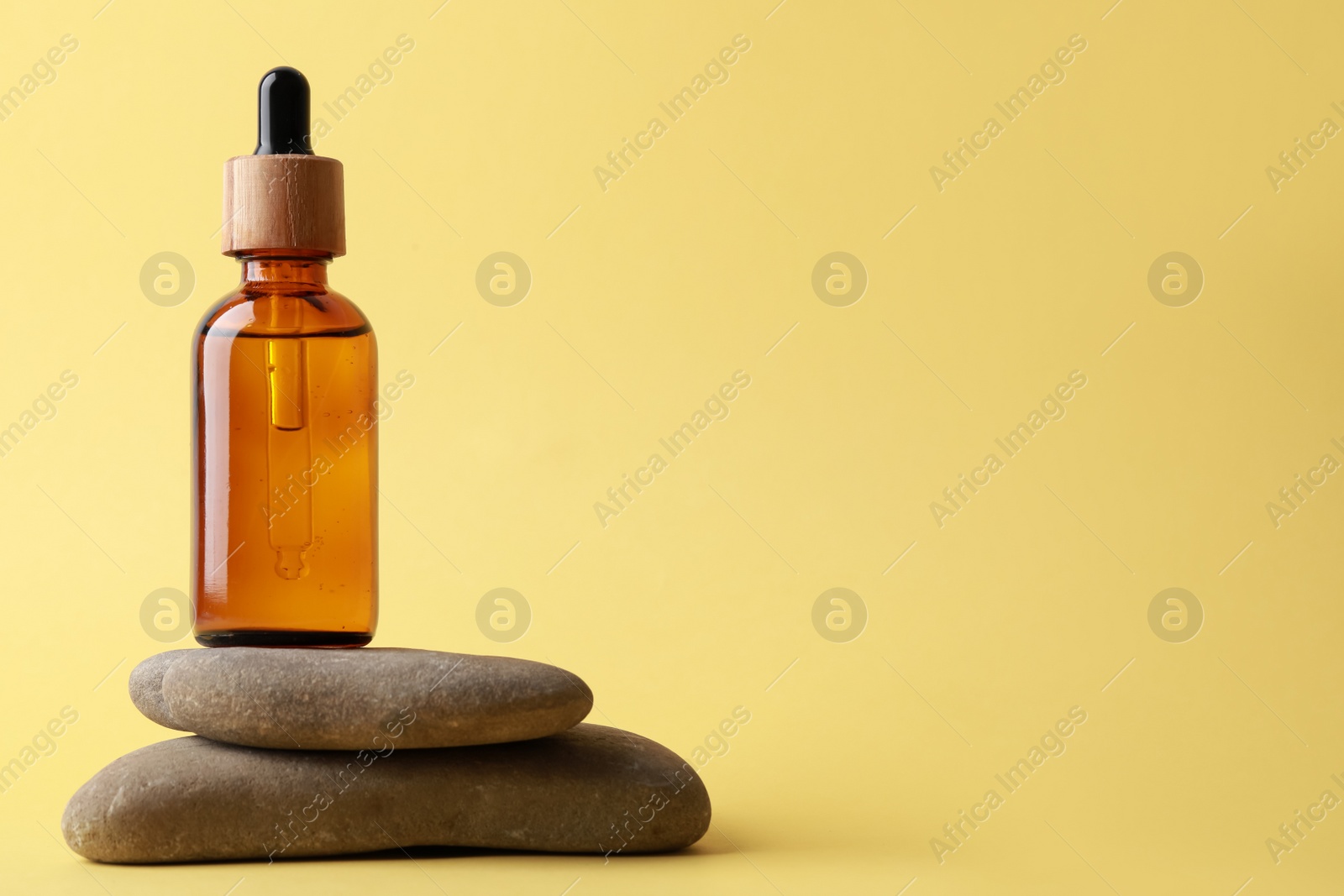 Photo of Bottle of face serum and spa stones on yellow background, space for text