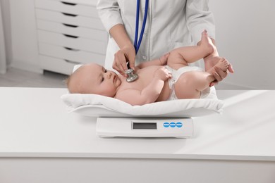 Pediatrician weighting and examining cute little baby with stethoscope in clinic, closeup