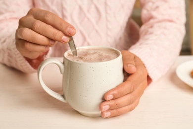 Woman holding cup of delicious cocoa drink at white table, closeup