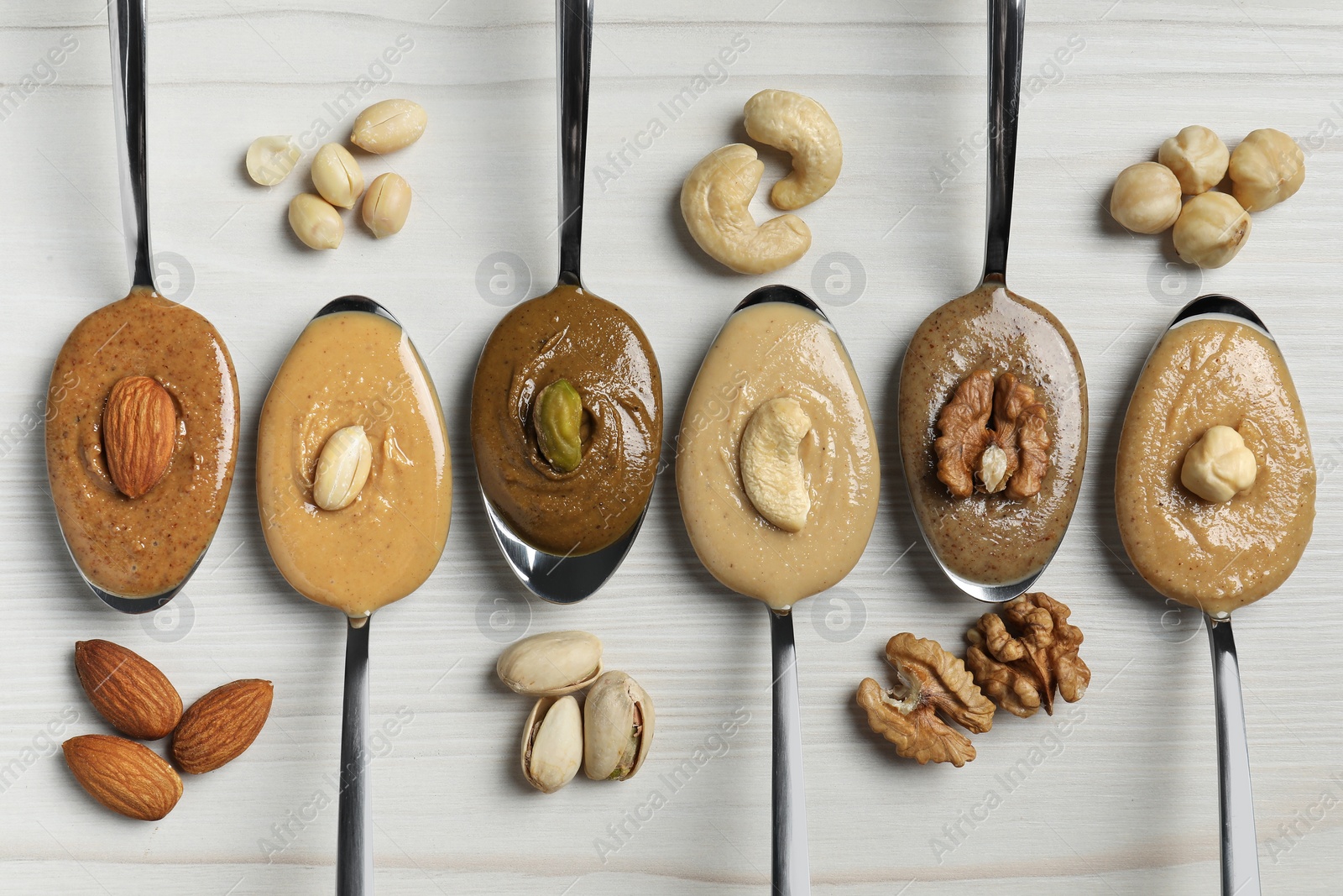 Photo of Tasty nut butters in spoons and raw nuts on white wooden table, flat lay