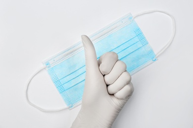 Doctor in medical gloves showing thumb up gesture near protective mask on white background, top view