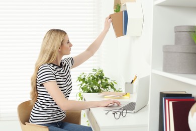 Photo of Home workplace. Woman taking book from shelf near laptop at white desk in room