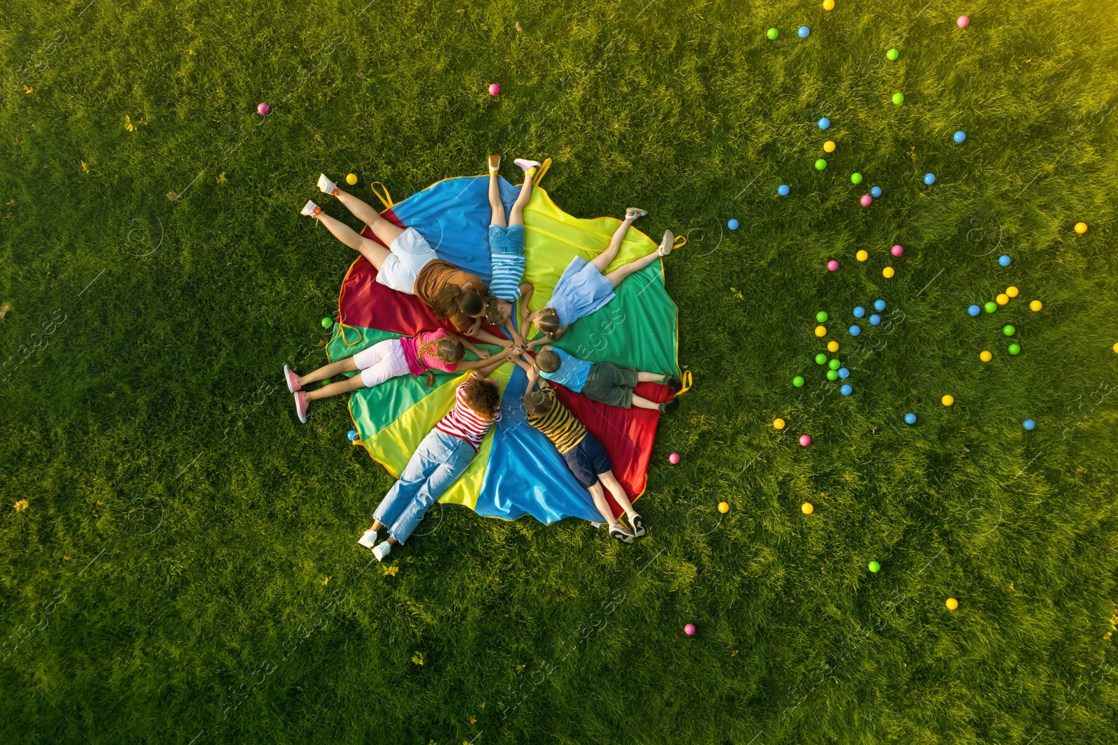 Image of Group of children with teachers holding hands together on rainbow playground parachute in park, top view. Summer camp activity