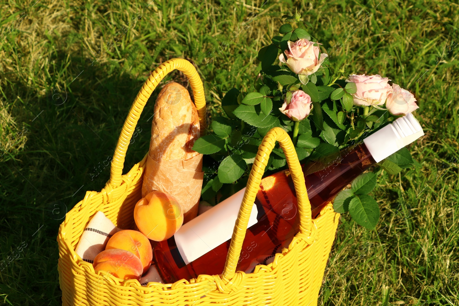 Photo of Yellow wicker bag with roses, peaches, baguette and wine on green grass outdoors