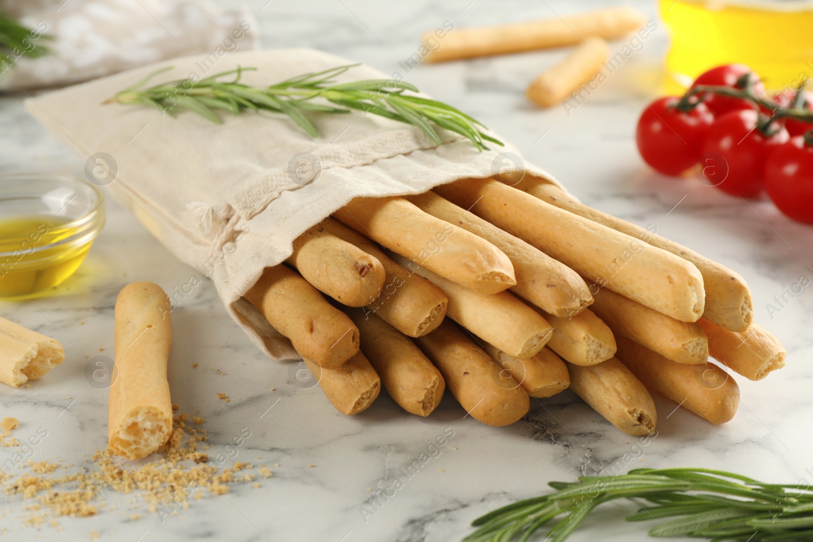 Photo of Delicious grissini sticks, oil, rosemary and tomatoes on white marble table, closeup