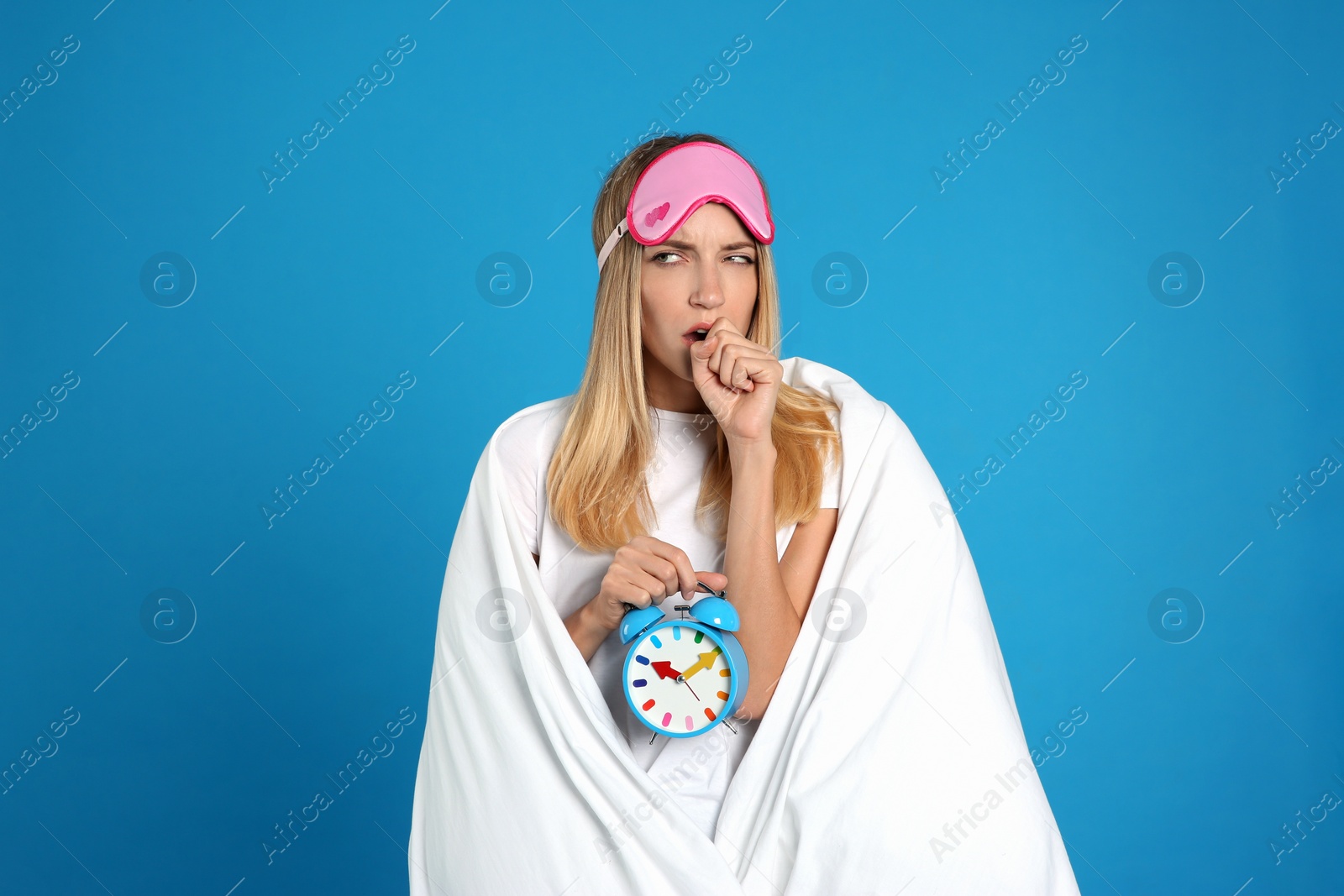 Photo of Young woman in sleeping mask wrapped with blanket holding alarm clock on blue background