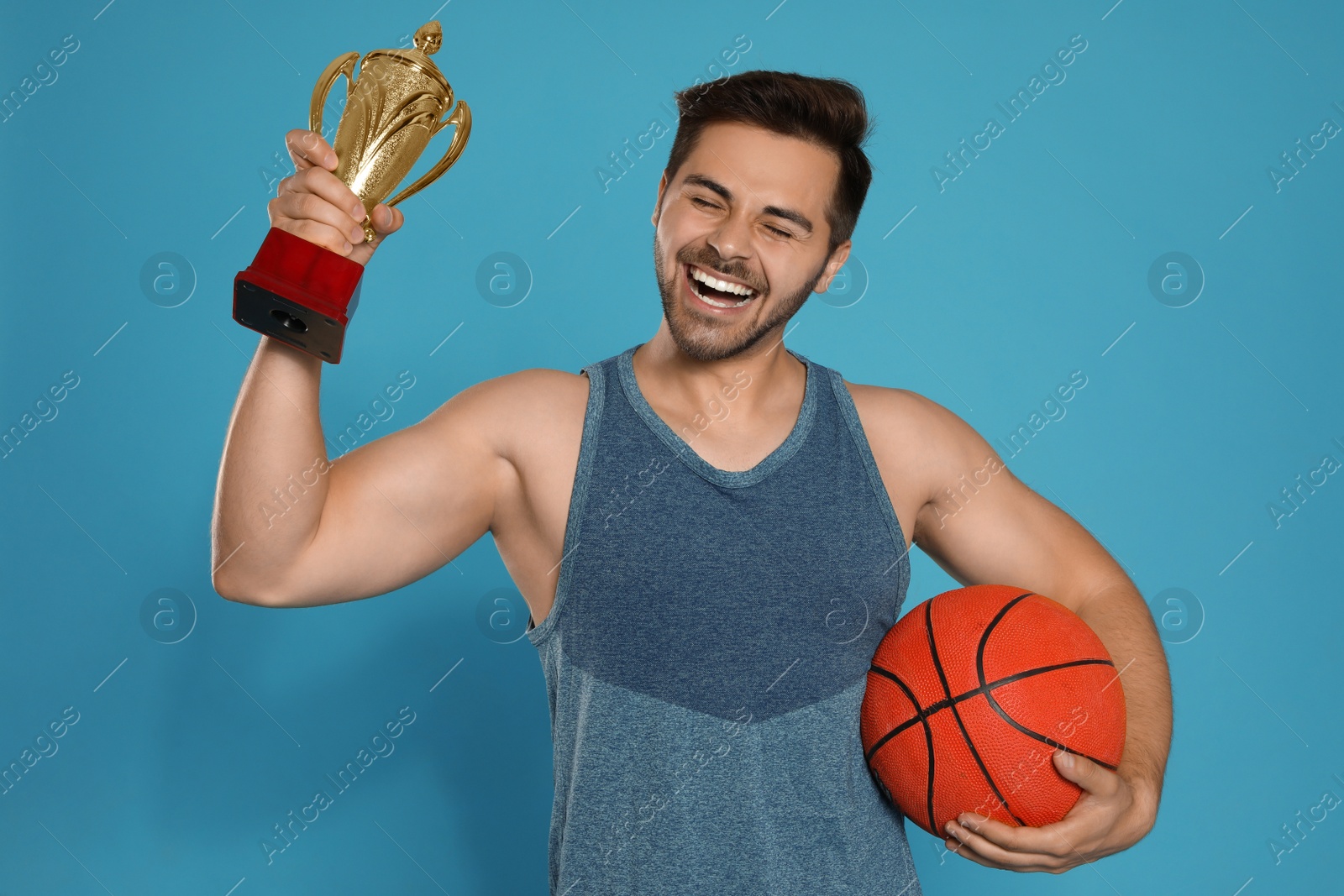 Photo of Portrait of happy young basketball player with gold trophy cup on blue background