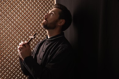 Photo of Catholic priest in cassock holding cross and praying to God in confessional, space for text