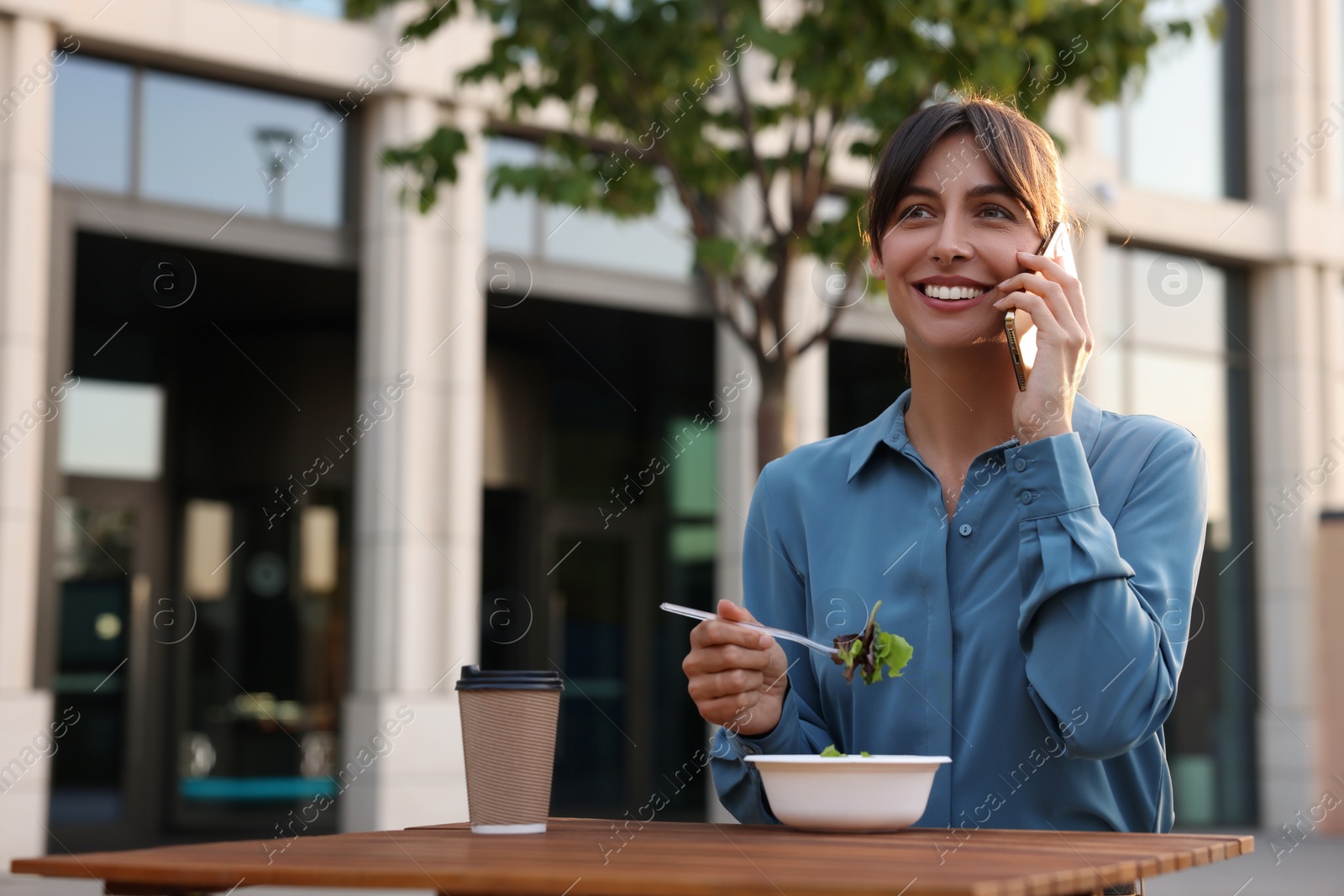 Photo of Happy businesswoman with plastic bowl of salad talking on smartphone during lunch at wooden table outdoors