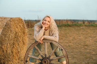 Photo of Beautiful woman posing with wooden cart wheel near hay bale in field. Autumn season