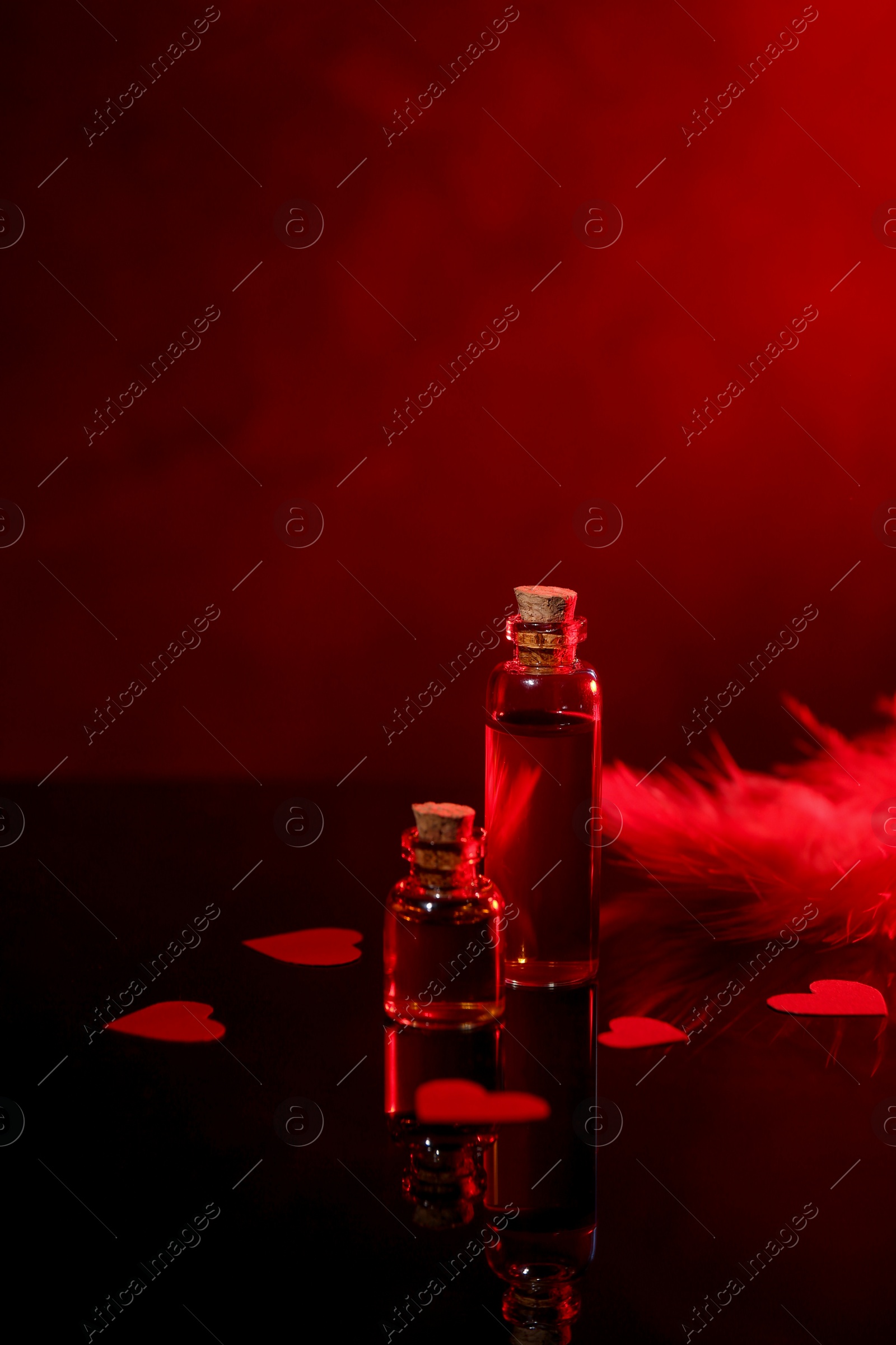 Photo of Bottles of love potion, paper hearts and feather on mirror surface against dark background, space for text. Red tone effect