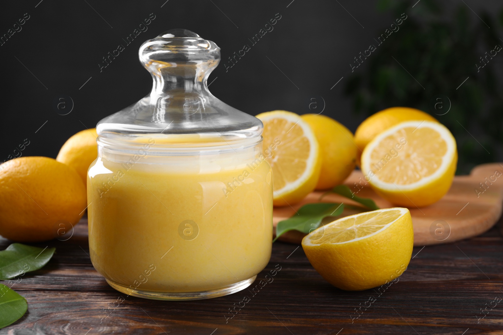 Photo of Delicious lemon curd in glass jar, fresh citrus fruits and green leaves on wooden table