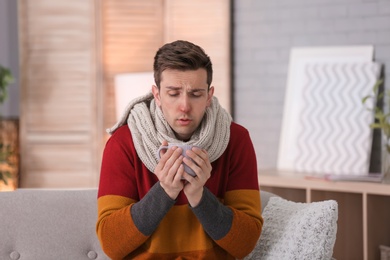 Sick young man with cup of hot drink suffering from cold on sofa at home