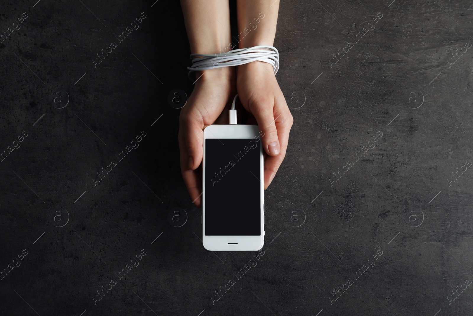 Photo of Woman holding mobile phone in hands tied with charging cable on grey background, above view. Loneliness concept