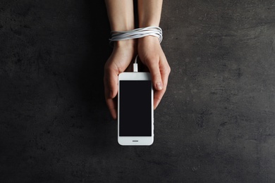 Woman holding mobile phone in hands tied with charging cable on grey background, above view. Loneliness concept