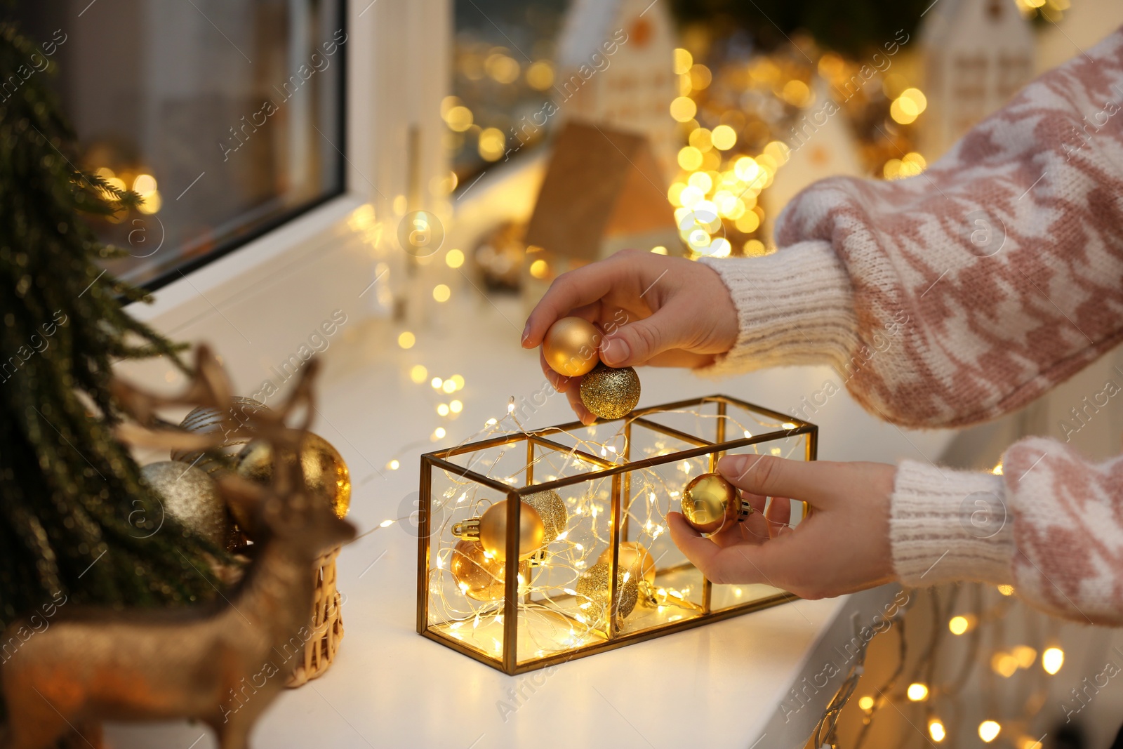 Photo of Christmas atmosphere. Woman putting beautiful baubles into decorative container on window sill indoors, closeup