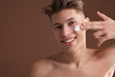 Handsome man applying moisturizing cream onto his face on brown background. Space for text