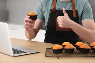 Photo of Man holding muffin near laptop and showing thumb up at table in kitchen, closeup. Time for hobby