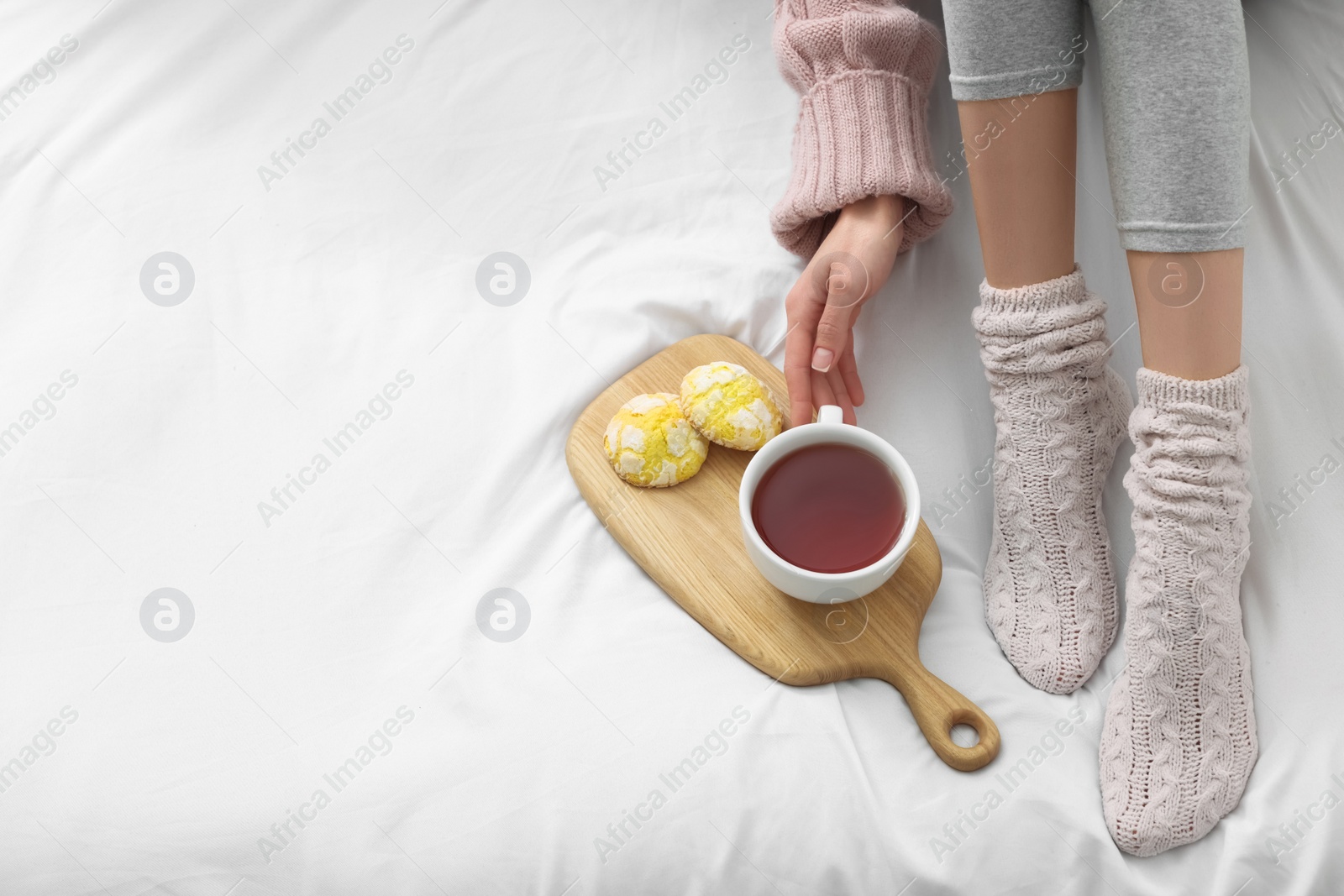 Photo of Woman with cup of tea and cookies wearing warm socks in comfortable bed, above view. Space for text