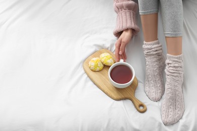 Woman with cup of tea and cookies wearing warm socks in comfortable bed, above view. Space for text