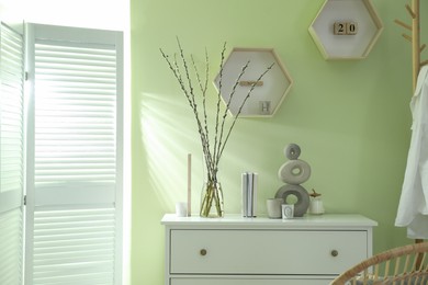Photo of Glass vase with pussy willow tree branches and decor on white chest of drawers near light green wall indoors