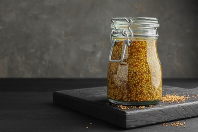 Photo of Whole grain mustard in jar and dry seeds on black table. Space for text