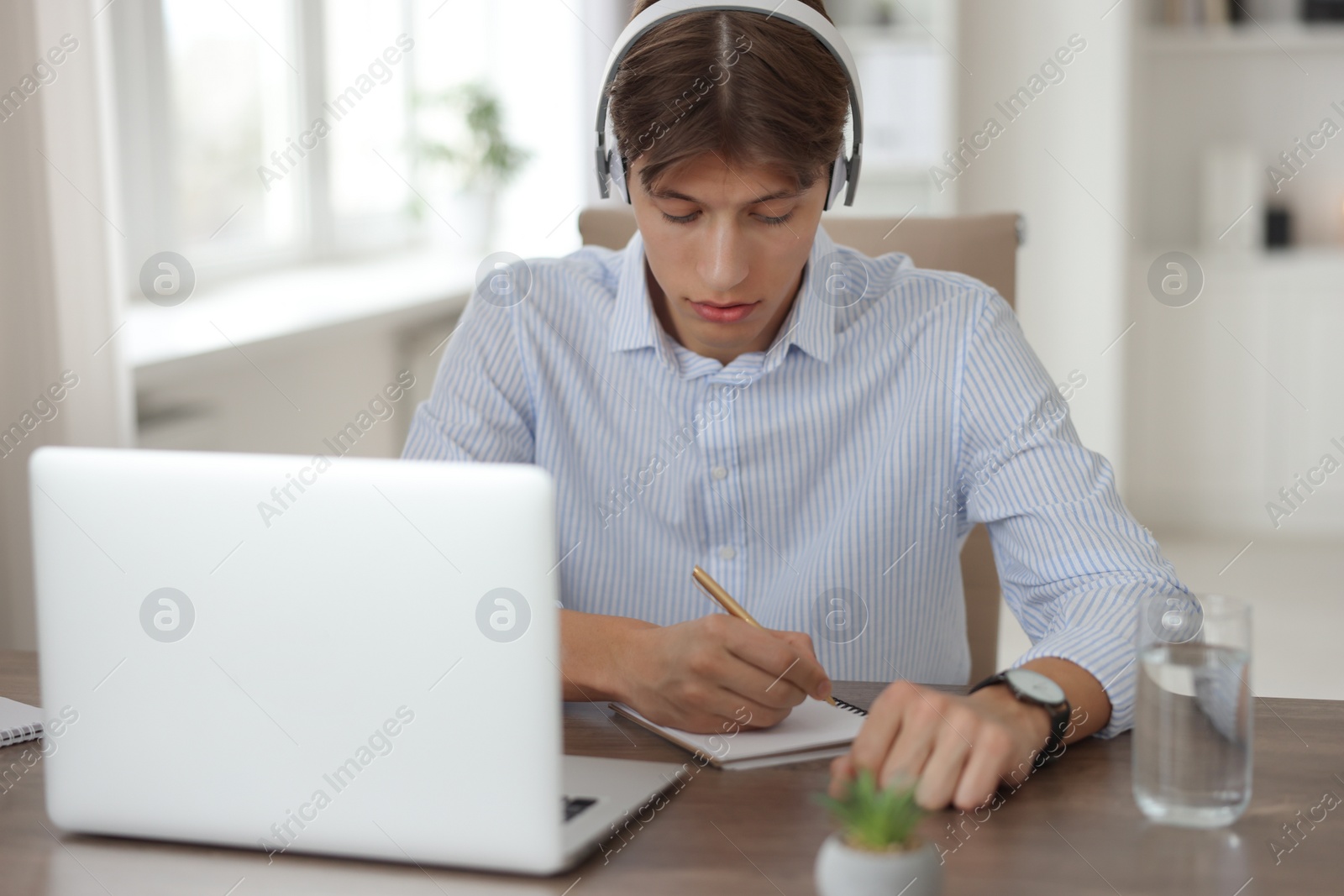 Photo of Man in headphones taking notes during webinar at wooden table in office