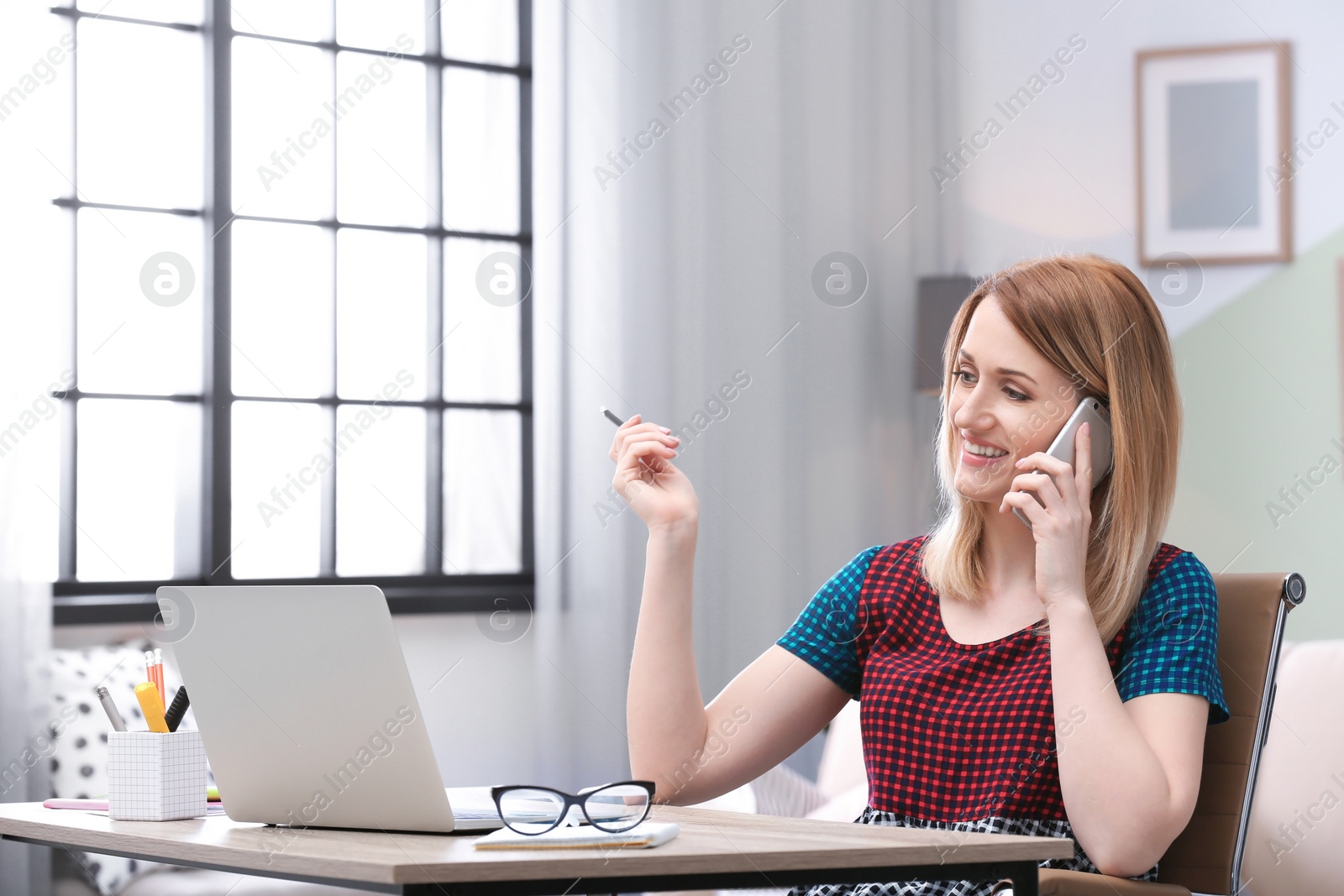 Photo of Young woman talking on phone while working with laptop at desk in home office