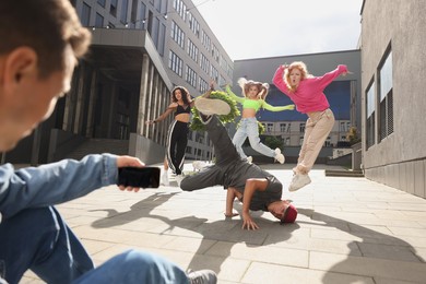 Photo of Group of people dancing hip hop while man recording them with smartphone outdoors, low angle view