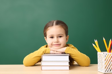 Happy little school child sitting at desk with books near chalkboard