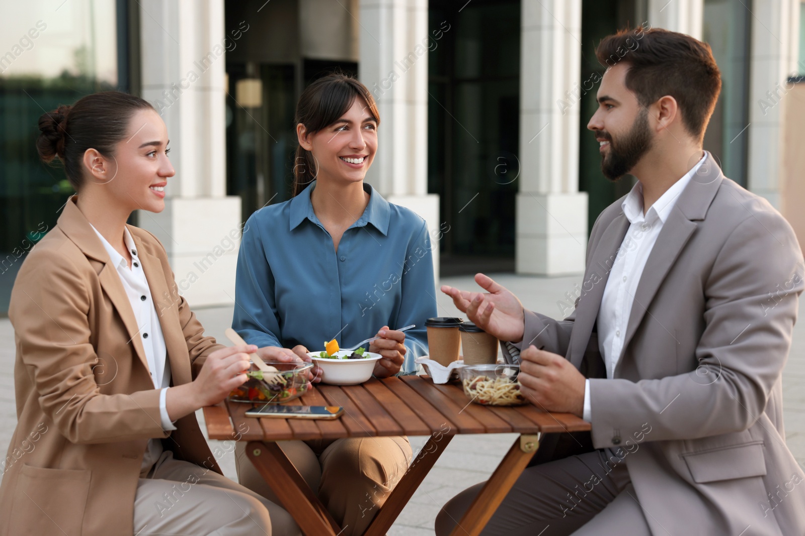 Photo of Business lunch. Happy colleagues spending time together at wooden table during break outdoors