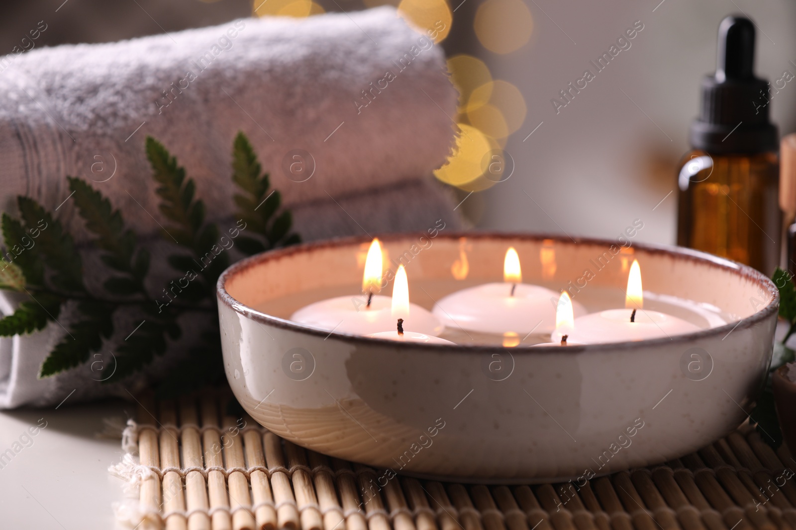 Photo of Spa composition. Burning candles in bowl, towels and bottle of essential oil on table against blurred lights, closeup