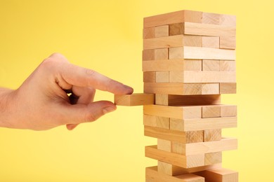 Photo of Playing Jenga. Man removing wooden block from tower on yellow background, closeup