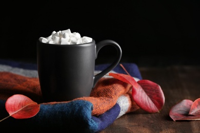 Cup of hot cozy drink with marshmallows, scarf and autumn leaves on table. Space for text