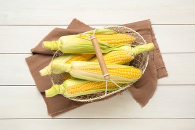 Photo of Tasty sweet corn cobs in metal basket on white wooden table, top view
