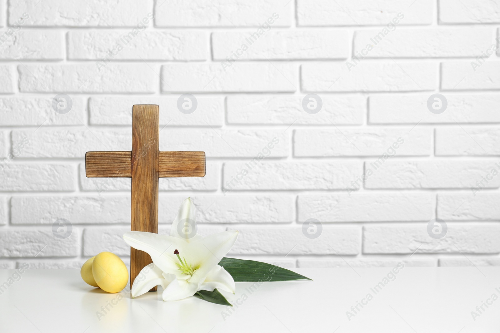 Photo of Wooden cross, Easter eggs and blossom lily on table against brick wall, space for text