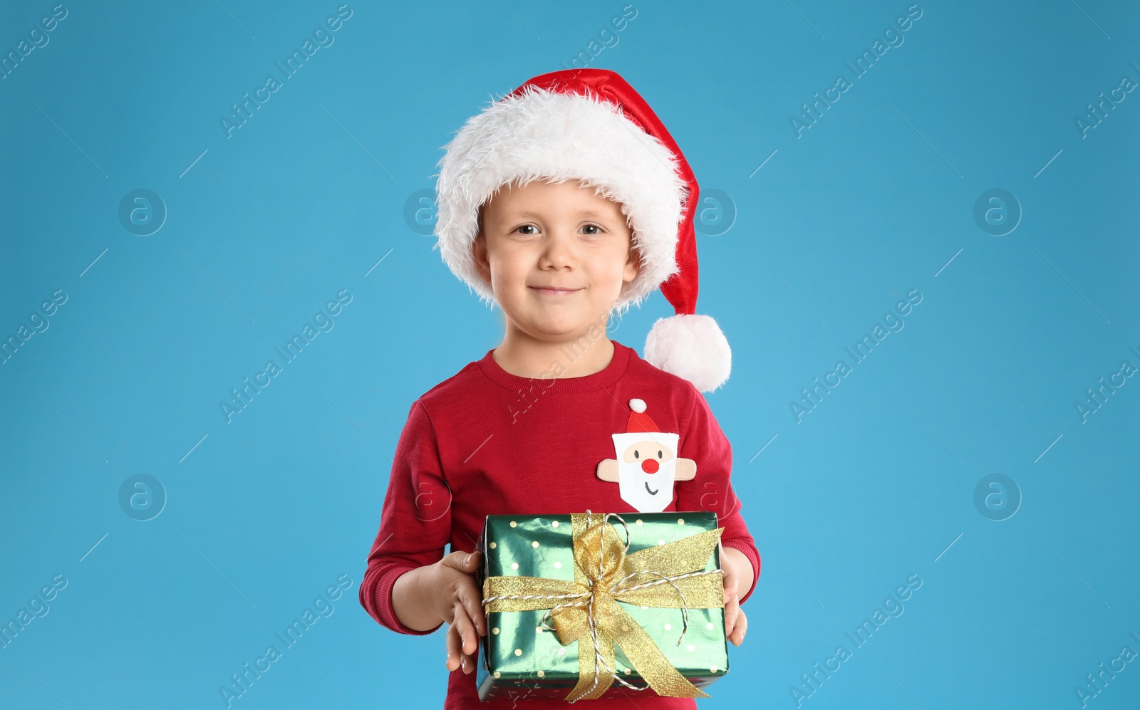 Photo of Cute child in Santa hat with Christmas gift on light blue background