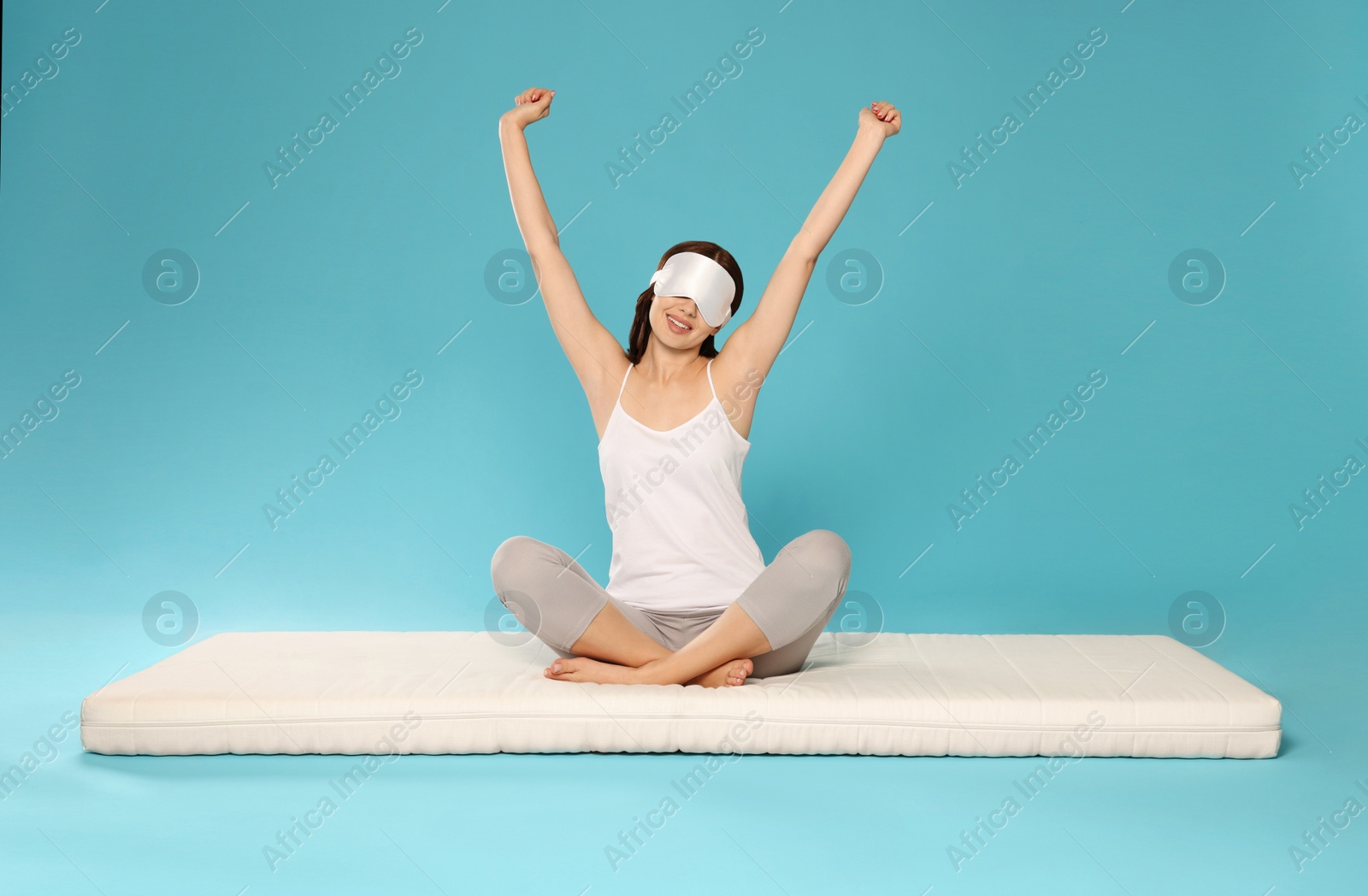 Photo of Woman in sleep mask stretching on soft mattress against light blue background