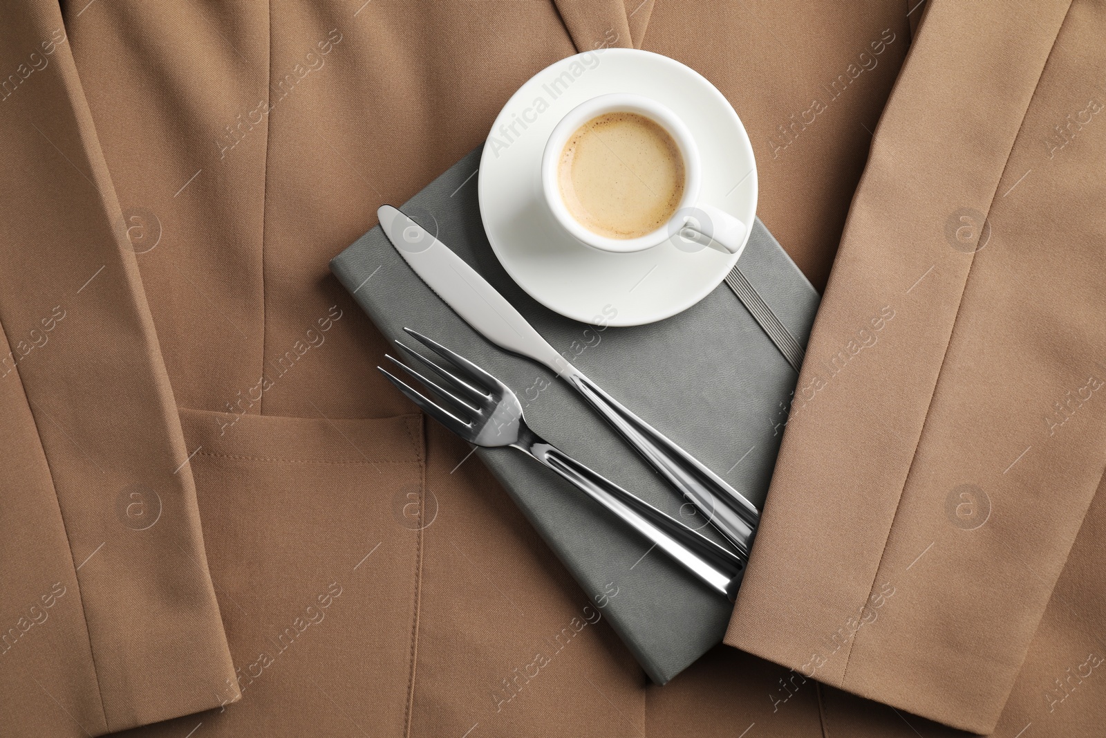 Photo of Business lunch concept. Cup of coffee, light gray notebook and cutlery on beige jacket, flat lay