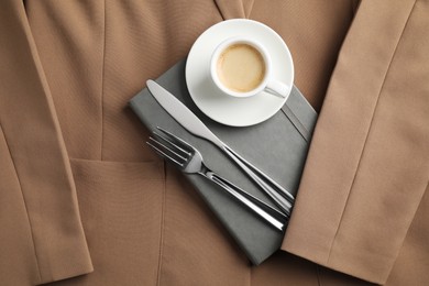 Photo of Business lunch concept. Cup of coffee, light gray notebook and cutlery on beige jacket, flat lay