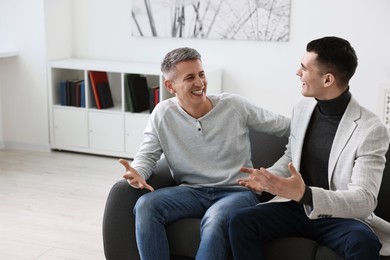 Photo of Happy men talking while sitting on sofa at home