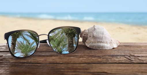 Palms reflecting in sunglasses on wooden desk with seashell at sandy beach. Banner design