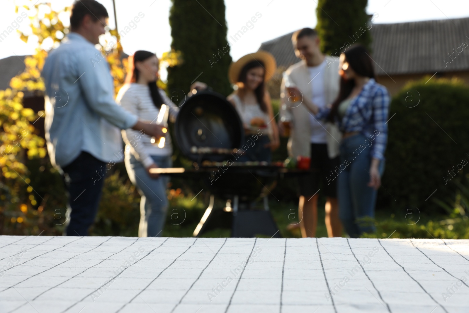 Photo of Empty white wooden table and blurred view of friends having barbecue party outdoors. Space for text