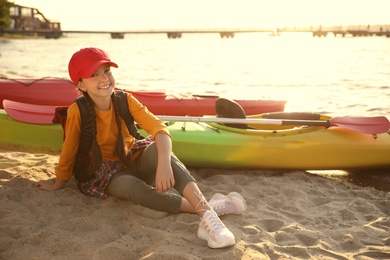 Photo of Happy girl sitting near kayak on river shore. Summer camp activity
