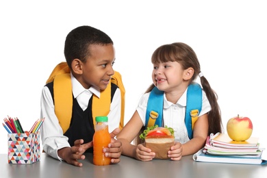 Schoolchildren with healthy food and backpacks sitting at table on white background