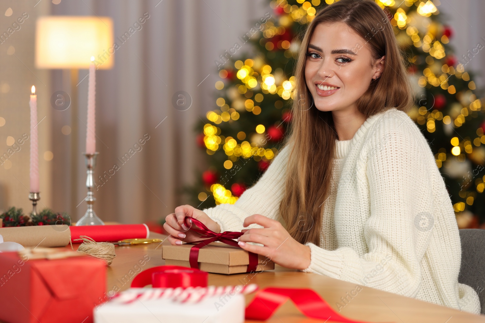 Photo of Beautiful young woman decorating Christmas gift at table in room